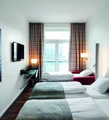 Modern hotel room with large window view. Features two beds with white and gray bedding, a wall-mounted TV, a patterned wall art, and a dark wood floor. Natural light illuminates the space, complemented by neutral decor.