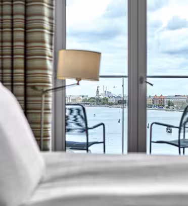 View from a hotel room with an unmade bed in the foreground, looking out through glass doors onto a balcony with two chairs. Beyond the balcony, theres a body of water, industrial buildings, and a cloudy sky in the distance.